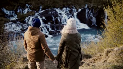 Vista-posterior-de-la-joven-pareja-elegante-levantando-las-manos-y-sentirse-feliz,-libertad-cerca-de-las-cascadas-de-Islandia