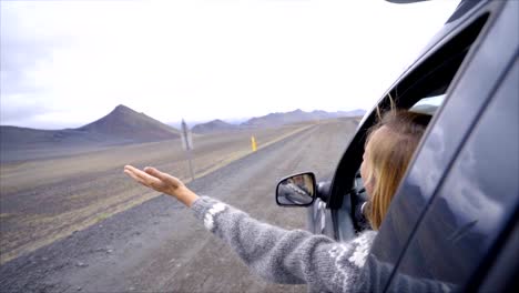 Cheerful-caucasian-female-inside-car-looking-at-road-arms-outstretched-,-mountain-volcanic-landscape.-Road-trip-concept.--Blond-hair-girl-looking-at-landscape.-SLOW-MOTION