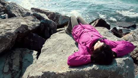 Young-Caucasian-woman-with-pink-coat-lying-on-the-rocks-near-the-wavy-sea