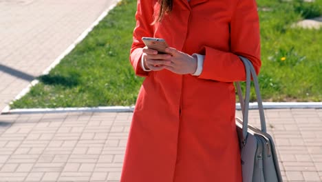 Unrecognizable-woman-in-red-coat-waits-for-someone-and-checks-her-phone,-texting.-Close-up-hands.