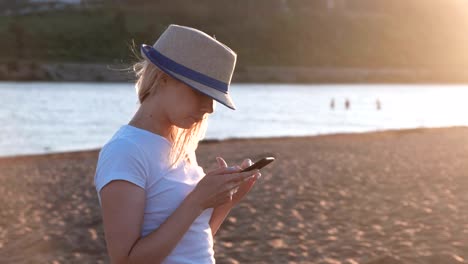 Beautiful-blonde-girl-in-a-hat-types-a-message-on-her-mobile-phone-on-the-beach-at-sunset.