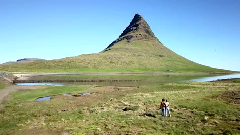 Drone-shot-aerial-view-of-caucasian-couple-contemplating-the-famous-Kirkjufell-mountain-peak,-nature-background-with-grass-and-mountains.-Shot-in-West-Iceland,-Springtime.-People-travel-lifestyles-concept--4K-video