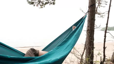 Young-caucasian-male-resting-lying-on-blue-hammock-outdoors-near-lake.