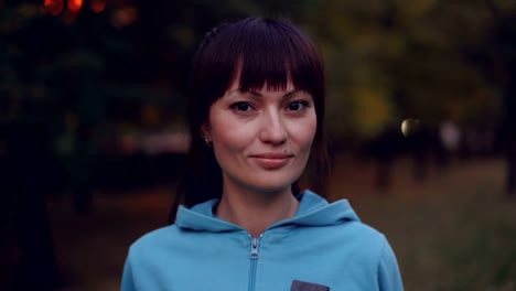 Retrato-de-hermosa-deportista-joven-con-pelo-largo-oscuro-y-ojos-marrones-mirando-a-cámara-y-sonrisa-permanente-al-aire-libre-en-otoño-noche-llevar-capucha.