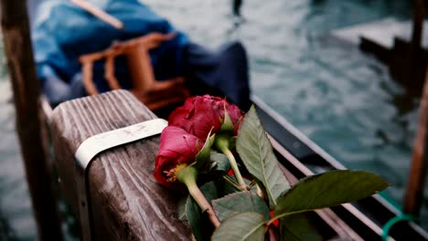 Close-up-shot-of-beautiful-red-roses-lying-on-wooden-canal-pier,-traditional-Italian-gondola-rocking-on-waves-nearby.