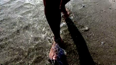 Men-barefoot-legs-silhouettes-walking-along-sea-shore-shallow-water-with-waves-splashing.-4k-trekking-steadicam--front-view-pov-shot