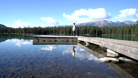 Young-woman-walks-on-wooden-pier-above-stunning-mountain-lake-scenery,-arms-wide-open