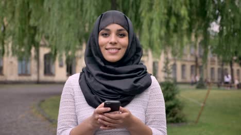 Young-beautiful-muslim-girl-in-hijab-is-typing-message-on-her-smartphone,-watching-at-camera-and-smiling-in-blue-weather,-communication-concept,-religious-concept