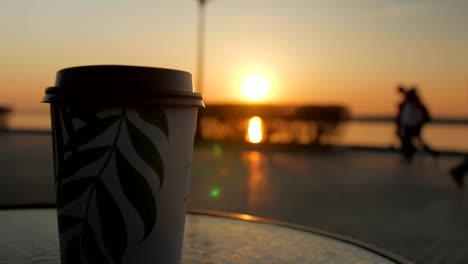 tazas-con-café-están-de-pie-sobre-una-mesa-en-el-fondo-de-puesta-de-sol,-mano-femenina-está-tomando-una