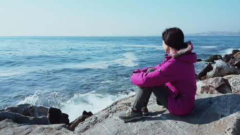 Young-Caucasian-woman-with-pink-coat-sitting-on-the-rocks-near-the-wavy-sea