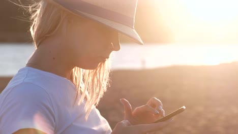 Beautiful-blonde-girl-in-a-hat-types-a-message-on-her-mobile-phone-on-the-beach-at-sunset.