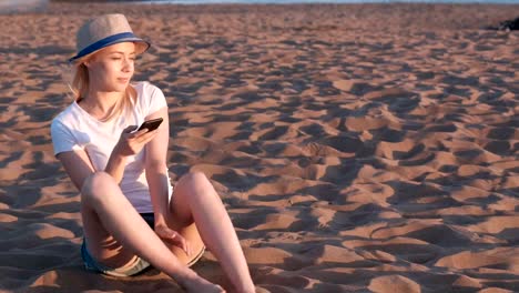 Schöne-blonde-Mädchen-mit-Hut-am-Strand-bei-Sonnenuntergang-mit-Handy-sitzen.