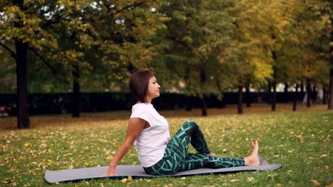 Hermosa-chica-en-ropa-de-deporte-es-ejercicio-en-el-Parque-practicando-giros-sentado-y-adelante-curvas-en-estera-de-yoga-cálido-día-de-otoño.-Concepto-de-naturaleza,-deportes-y-salud.