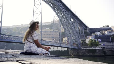 Dame-mit-Hut-aus-der-Tasse-trinken-und-sitzen-am-Ufer-in-der-Nähe-von-Brücke