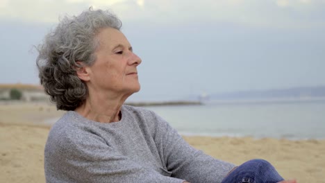 Thoughtful-senior-lady-sitting-on-beach-and-looking-at-distance.