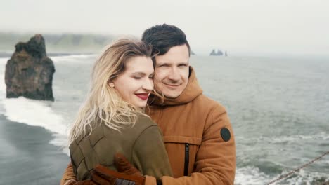 Portrait-of-young-happy-couple-standing-on-the-black-beach,-near-the-troll-toes-rocks-in-Iceland-and-hugging-together