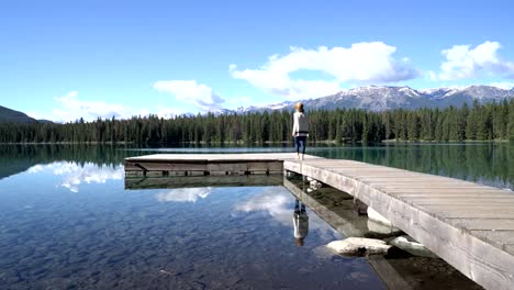 Young-woman-walks-on-wooden-pier-above-stunning-mountain-lake-scenery,-arms-wide-open