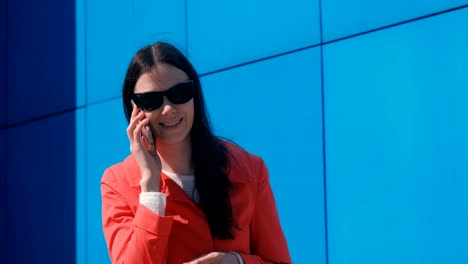 Mujer-morena-joven-en-gafas-de-sol-habla-en-el-teléfono-al-lado-del-edificio-azul.