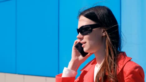 Portrait-of-young-brunette-woman-in-sunglasses-speaks-on-the-phone-beside-blue-building.