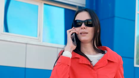 Portrait-of-young-brunette-woman-in-sunglasses-and-red-coat-speaks-on-the-phone-beside-blue-building.