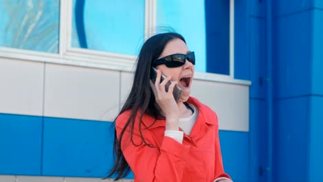 Portrait-of-young-brunette-woman-in-sunglasses-and-red-coat-speaks-on-the-phone-beside-blue-building.