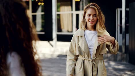 Attractive-girl-is-talking-to-her-friend-standing-in-the-street-in-the-city-and-gesturing-sharing-news.-Emotional-young-woman-with-blond-hair-is-wearing-trendy-coat.