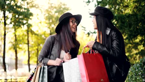 Excited-girl-is-looking-at-her-friend's-purchases-in-bags,-asking-questions-about-prices-and-collections-and-expressing-excitement-about-shopping-standing-in-the-street-in-city.