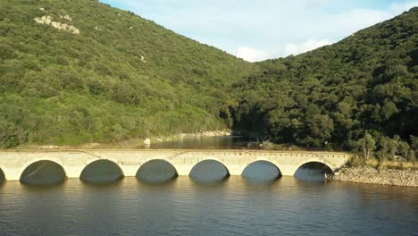 Video-from-above,-aerial-view-of-a-Roman-bridge-on-a-beautiful-lake-sorrounded-by-some-green-hills-in-Sardinia,-Italy.