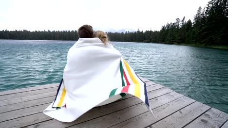 Young-couple-sharing-a-romantic-moment-in-nature,-wrapped-in-blanket-on-lake-pier,-Canada