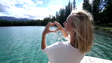 Young-woman-making-heart-shape-finger-frame-on-mountain-lake