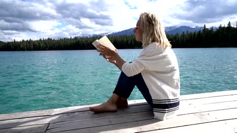 Young-woman-relaxing-on-lake-pier-with-book,-Canada