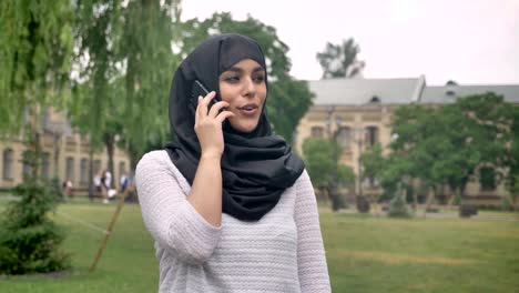 Young-muslim-woman-in-hijab-is-talking-on-phone-in-blue-weather-in-park,-building-on-background,-religiuos-concept,-communication-concept