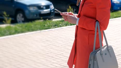 Nicht-erkennbare-Frau-im-roten-Mantel-wartet-auf-jemanden-und-überprüft-ihr-Handy-SMS.-Close-up-Hände.-Seitenansicht.