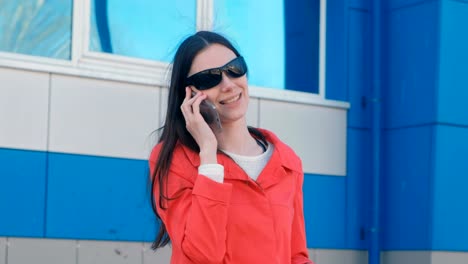 Portrait-of-young-brunette-woman-in-sunglasses-and-red-coat-speaks-on-the-phone-beside-blue-building.