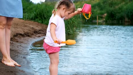 Kleinkind-spielt-im-Wasser-an-einem-Strand-am-Fluss-mit-einer-Schaufel-und-Topf-im-Sommer.-Mama-kümmert-sich-um-ihre-Tochter.