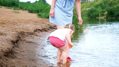 Small-child-is-playing-in-the-river,-the-girl-pours-water-out-of-the-pot-in-the-summer.-Mom-looks-after-her-daughter.