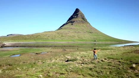 Vista-aérea-de-Drone-disparó-de-brazos-hombres-caucásico-levantados-para-las-emociones-positivas,-montaña-Kirkjufell-sobre-fondo.-Rodada-en-Islandia-occidental,-primavera.-Concepto-de-estilos-de-vida-sin-preocupaciones-la-gente-viaja