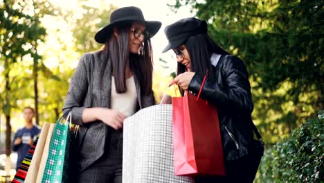 Happy-Asian-girl-with-shopping-bags-is-talking-to-her-friend-standing-in-the-street-then-showing-her-purchases-while-Caucasian-young-woman-is-expressing-positive-emotons.