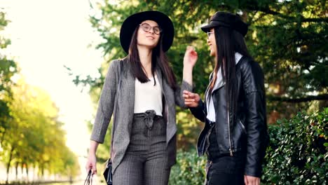 Chicas-felices-charlando-y-riendo-de-pie-al-aire-libre-en-acera-sosteniendo-bolsas-brillantes-discutiendo-día-en-centro-comercial.-Las-mujeres-están-usando-gafas-y-ropa-elegante.