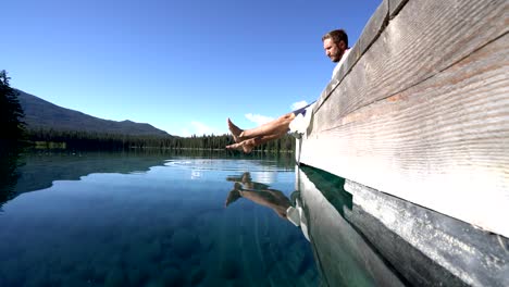 Man-sitting-on-wooden-pier-bay-stunning-lake-touches-the-water-surface-with-his-feet