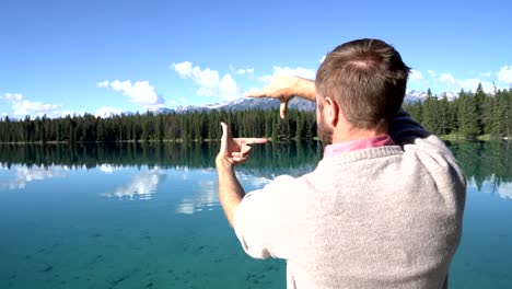 Man-stands-on-wooden-pier-above-stunning-lake-and-makes-a-frame-with-his-hands