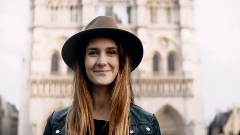 Portrait-of-young-beautiful-woman-in-hat-near-the-Notre-Dame-in-Paris,-France.-Female-looking-at-camera-and-smiling