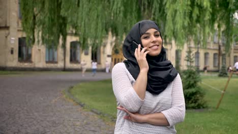 Young-muslim-woman-in-hijab-is-talking-on-phone-and-smiling-in-blue-weather,-building-on-background,-religiuos-concept,-communication-concept