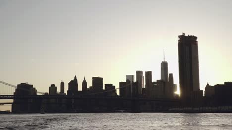 Una-puesta-de-sol-sunflare-brilla-a-través-de-skyline-de-Lower-Manhattan-y-el-puente-de-Brooklyn-en-Nueva-York,-Estados-Unidos-filmado-desde-el-East-River