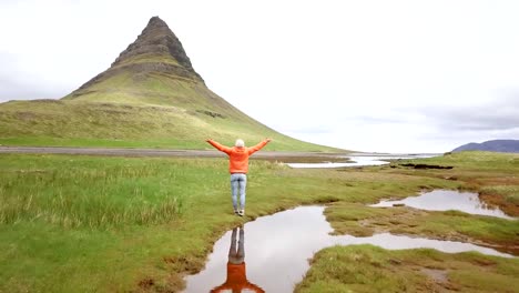 Drone-view-aerial-of-Young-woman-in-Iceland-arms-outstretched-for-freedom-Springtime-overcast-sky-at-famous-Kirkjufell-mountain--4K-footage