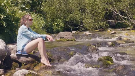 Mujer-turista-sentado-en-la-orilla-del-río-pedregoso-y-descansando-mientras-caminata-de-verano