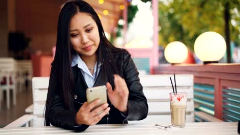 Lächelnde-asiatische-Studentin-ist-ruhen-im-Café-im-freien-und-mit-Smartphone-Bildschirm-zu-berühren-und-zu-beobachten.-Social-Media,-moderne-Technologie-und-City-Konzept.