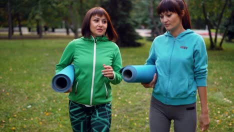 Cheerful-young-women-are-walking-on-grass-and-talking-holding-yoga-mats-after-practice-in-city-park.-Communication,-healthy-lifestyle-and-friendship-concept.