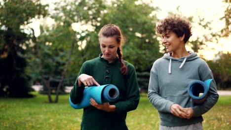 Hübsche-Mädchen-gehen-im-Park-hält-Yoga-Matten-und-reden-über-Sportgeräte,-Lächeln-und-lachen.-Erholung,-aktiven-Lebensstil-und-Natur-Konzept.