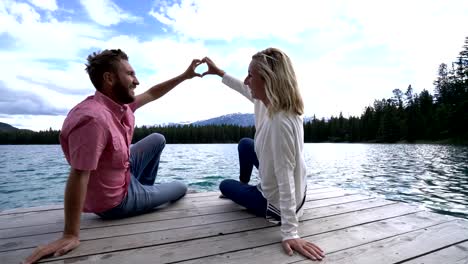 Joven-pareja-en-el-muelle-del-lago-haciendo-corazón-forma-dedo-marco